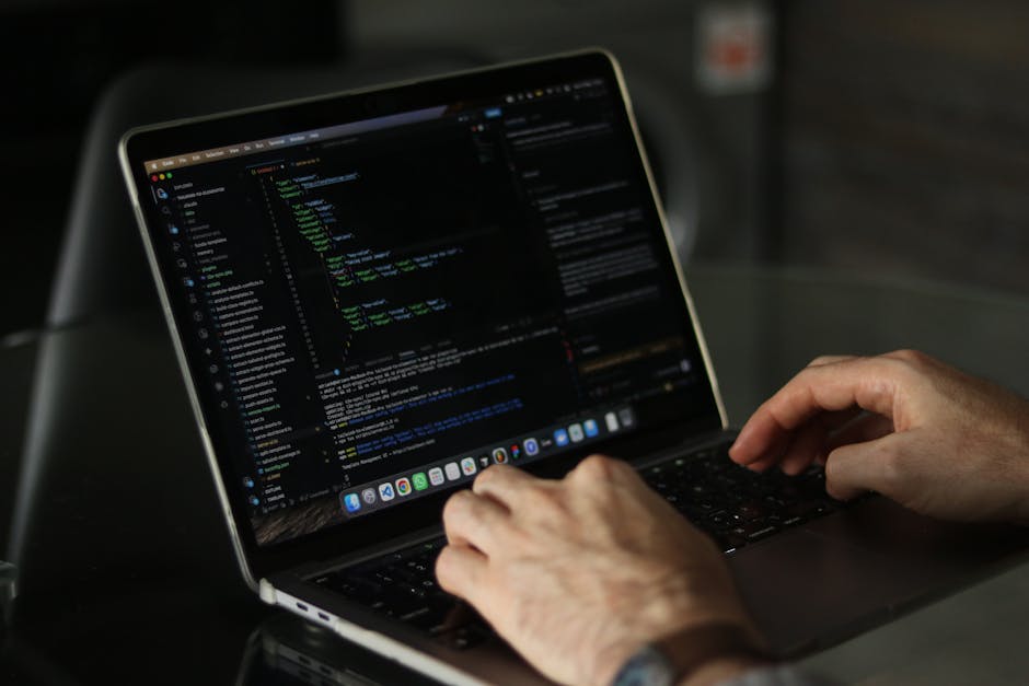 A close-up view of a person's hands working on a laptop, which displays a dark-themed coding interface with multicolored lines of code and text buffers. The laptop is placed on a dark, reflective surf